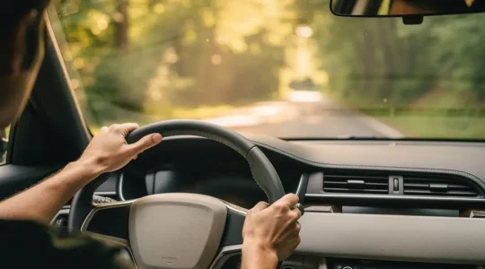 Vue intérieure d'une voiture avec les mains d'un conducteur sur le volant, baignée de lumière naturelle, symbolisant une conduite sereine et économique.